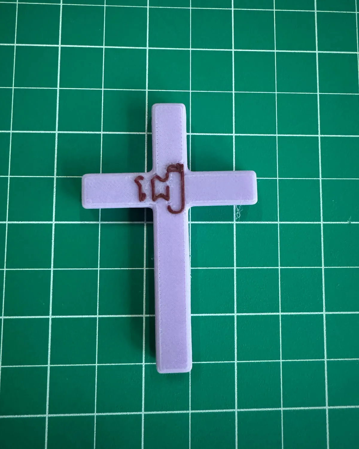 A lavender 3D-printed cross with the Khmer word for "believe" in raised letters at the intersection